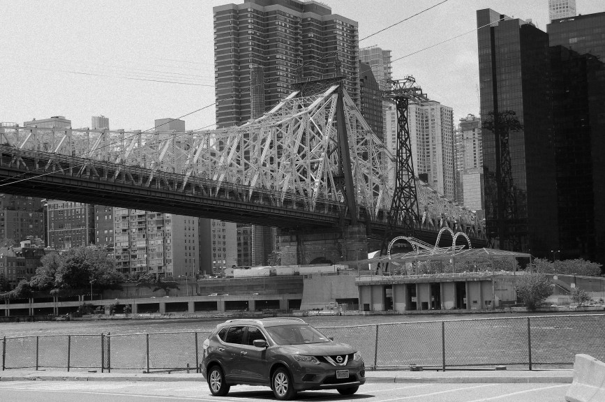 Black and white photo of a red car on Roosevelt Island with the Queensboro Bridge and Manhattan in the background.  Taken by N.A. Ferrell on June 8, 2019.