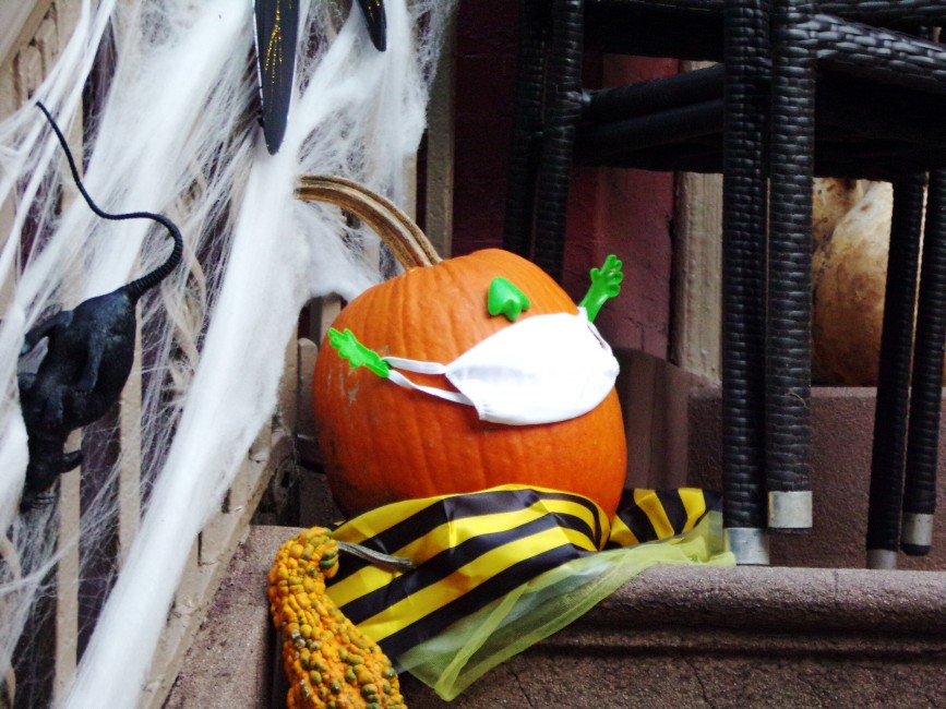 A pumpkin on a stoop wearing a protecting cloth mask, affixed to its face by two plastic hands, for Halloween.