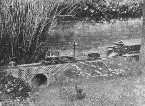 1895 photo of a model train leaving a model train station and crossing a miniature skew bridge.