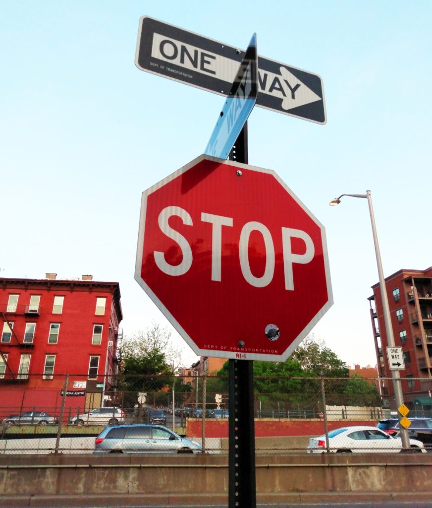 A combination stop/one way sign in Columbia Street Waterfront District with the BQE and Carroll Gardens, Brooklyn, in the background.