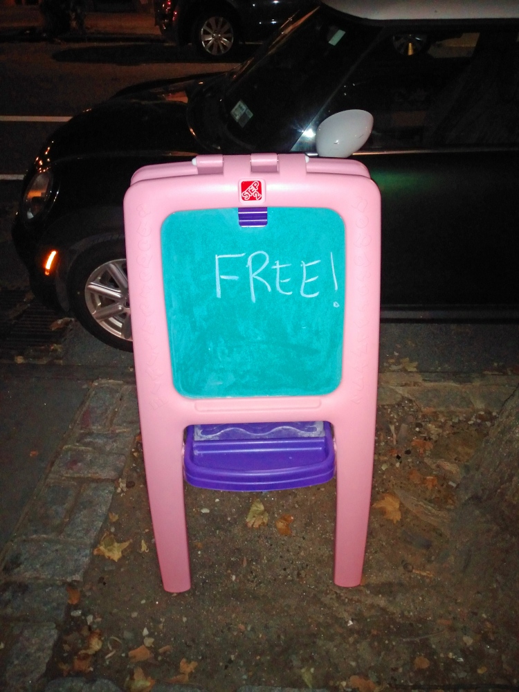 A chalkboard built into a plastic pink easel sitting in a tree pit in Cobble Hill, Brooklyn.  "FREE!" is written on the chalkboard in chalk, indicating that it was there for the taking.
