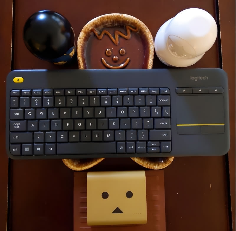A Logitech media keyboard sitting atop a Gingerbread Man plate. Next to the Gingerbread Man is a Kokeshi doll, a nesting doll, and a Danbo charger.