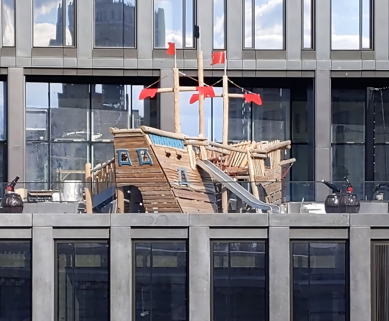 Photo of a playground in the shape of a boat on the balcony of a DUMBO Brooklyn apartment building, seen from the Brooklyn Bridge.  Balanced view showing front and back.