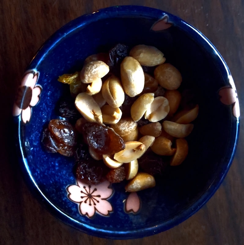 A photo of a small dark blue Japanese-style rice bowl with peanuts and black and yellow raisins on it. You can see a pink sakura flower painted on the inside of the bowl. The bowl sits on a dark wood surface. Photo taken and edited by N.A. Ferrell.