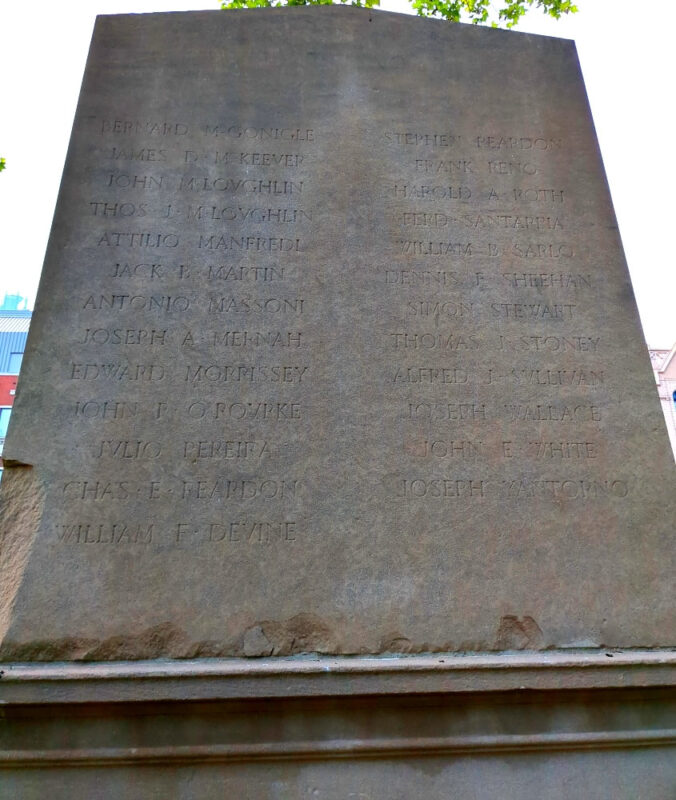 Photograph of the back of the McLaughlin Park World War I Memorial at the intersection of Tillary and Jay Streets in Downtown Brooklyn. A 12-foot stele sits on a 15 inch base. The top of the stele lists the names of 23 U.S. soldiers who died in World War I.