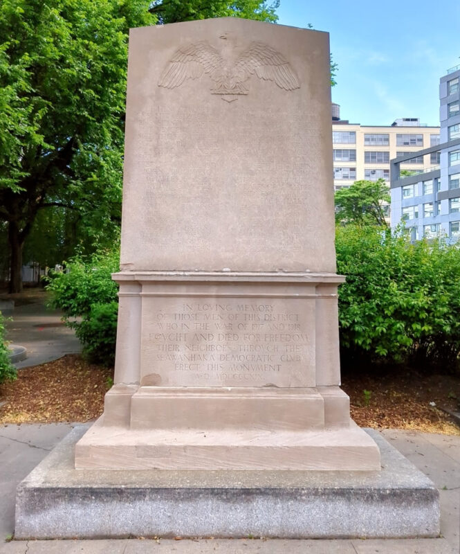 Photograph of the front of the McLaughlin Park World War I Memorial at the intersection of Tillary and Jay Streets in Downtown Brooklyn. A 12-foot stele sits on a 15 inch base. The top of the stele lists the names of 42 U.S. soldiers who died in World War I and the bottom features the dedication.
