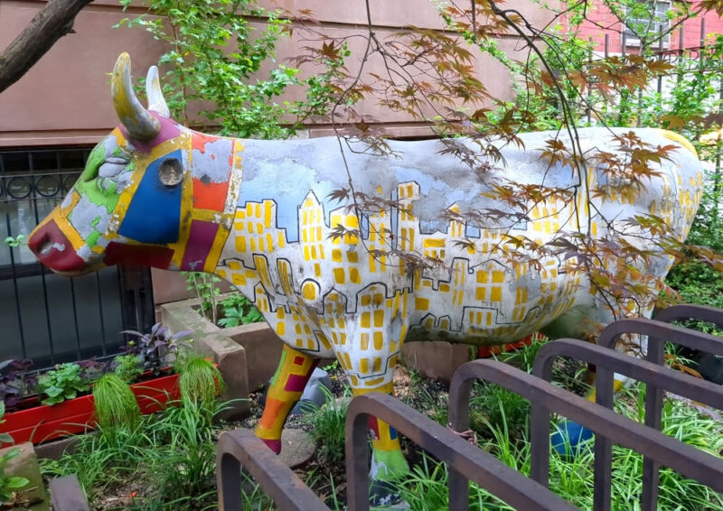 A cow statute in a small front yard in lower Manhattan. The cow statue is about 6-7 feet long and has horns. Its torso is painted witha  white and yellow sky line under a bluew cloud. Its face has patches of green, blue, maroon, and orange.