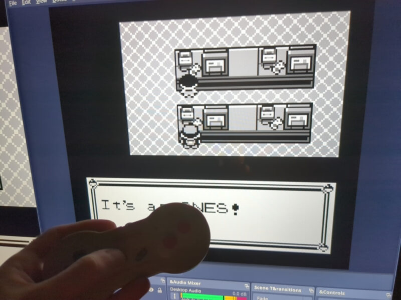 My hand holds an authentic NES 2 controller connected to my computer with an NES-to-USB adapter in front of my TV, on which I am playing Pokemon Red with the Gambatte emulator. In Pokemon Red, I am standing in front of an in-game SNES in the Celadon City Department Store. The on-screen dialogue reads "It's an SNES!" but I am holding the controller to block the n and S so it looks like it reads It's a NES!"