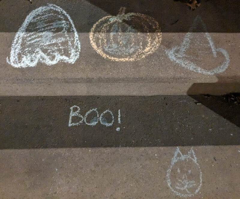 Halloween themed chalk art on a stoop. On the top step from left to right there is a Pacman-like ghost, a detailed pumpkin, and a witch's hat. On the next step down there is the text "BOO!" and a round cat face.