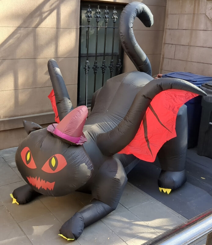 A large inflatable cartoonish black cat with a purple hat and bat wings, crouched in an aggressive pose, seen in front of a brownstone.