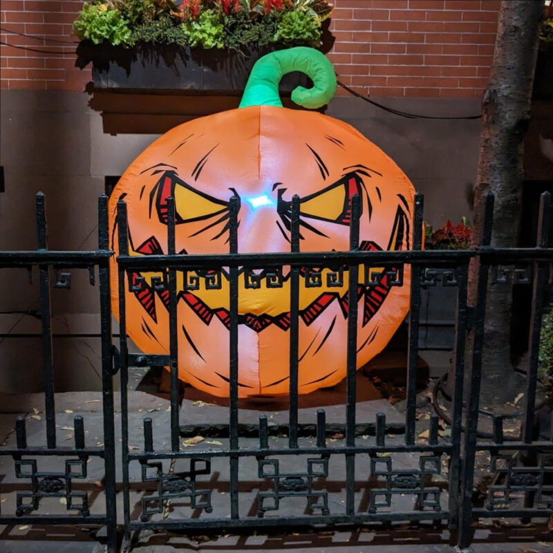 An inflatable Jack-o'-Lantern behind a fence in front of a brick house.