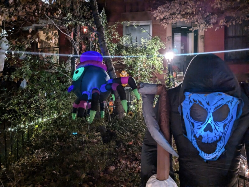 In inflatable grim reaper in the foreground with a large inflatable spider, a small cloth ghost, and a beam of light from a lantern in front of homes in the background.