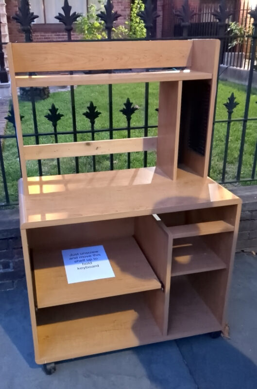 Front view of free children's desk with rollers left outside a home in Carroll Gardens. A note on one of the bottom shelves reads "Just unscrew and move this shelf up to hold keyboard"