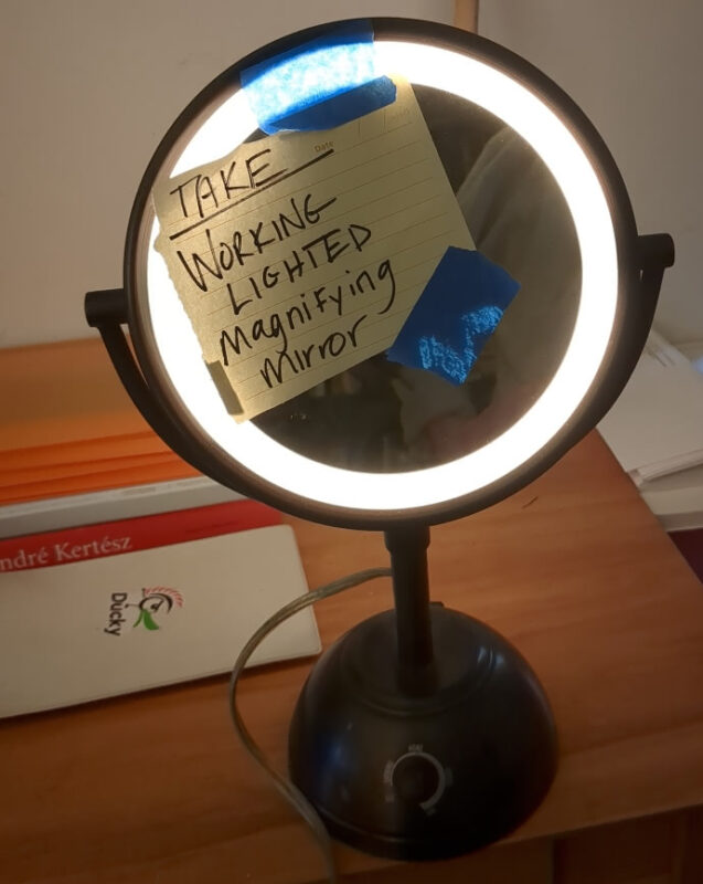 A lighted magnifying mirror on a desk. A note taped to its front reads "TAKE WORKING LIGHTED Magnifying mirror"