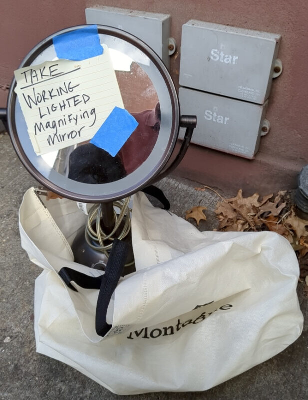 A magnifying mirror sticking out of a reusable shopping bag on the sidewalk in Brooklyn Heights. A note taped to its front reads "TAKE WORKING LIGHTED Magnifying mirror"