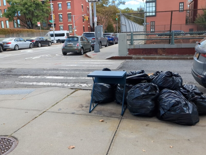 Blue desk with z-shaped legs next to a pile of garbage bags on curb in Columbia Street Waterfront District. In the background is the Congress Street crossing over the BQE and the Cobble Hill side of Hicks Street.