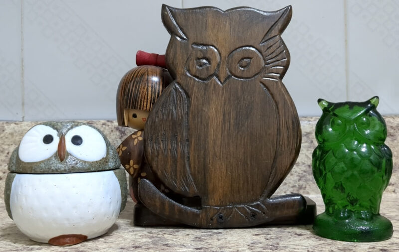 From lerft to right, a short brown and white owl with a Japanese aesthetic which is a condiment container, a tall dark wooden brown owl, and a green glass owl. There is a brown kokeshi doll peaking from behind the large owl.