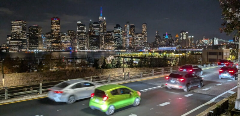 Photograph of the BQE at night with cars speeding along and the Manhattan skyline in the background taken from Grace Court Alley in Brooklyn Heights.