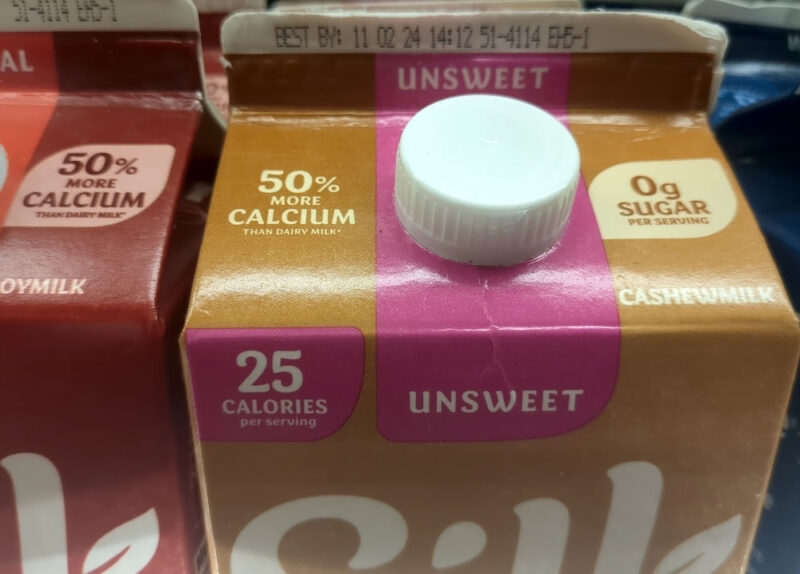 A carton of Silk cashew milk amongst other cartons of Silk non-dairy milk at the grocery store. Its sell-by date is 11/02/24, which was a problem since this photo was taken on December 15, 2024.