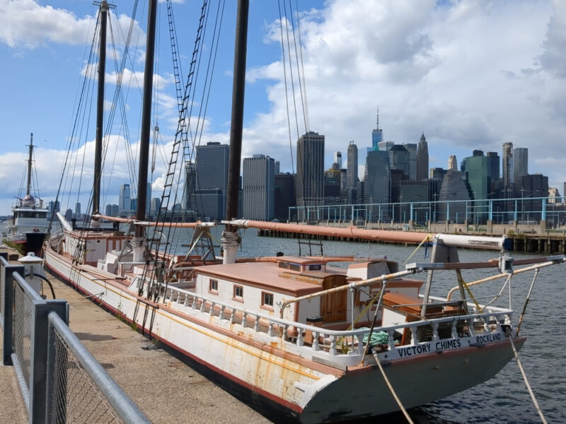 Full length photograph from behind of the Victory Chimes schooner docked at Brooklyn Bridge Park Pier 6. A small fireboat named the Gov. Alfred E. Smith is docked just in front of Victory Chimes. The Manhattan skyline is seen in the background.