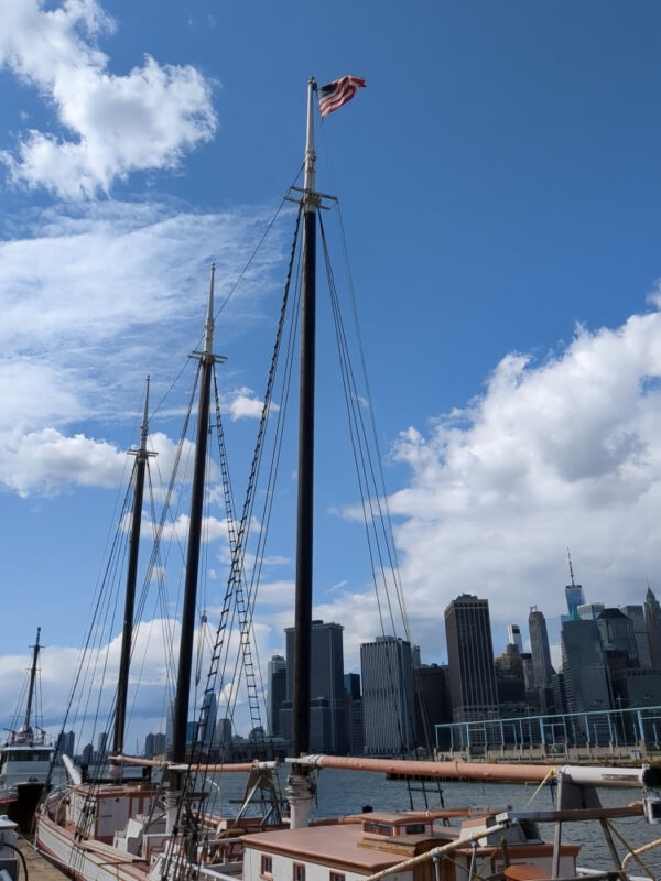 Full shot of the three wooden masts of the Victory Chimes as the ship is docked at Brooklyn Bridge Park. The Manhattan skyline is seen in the background.