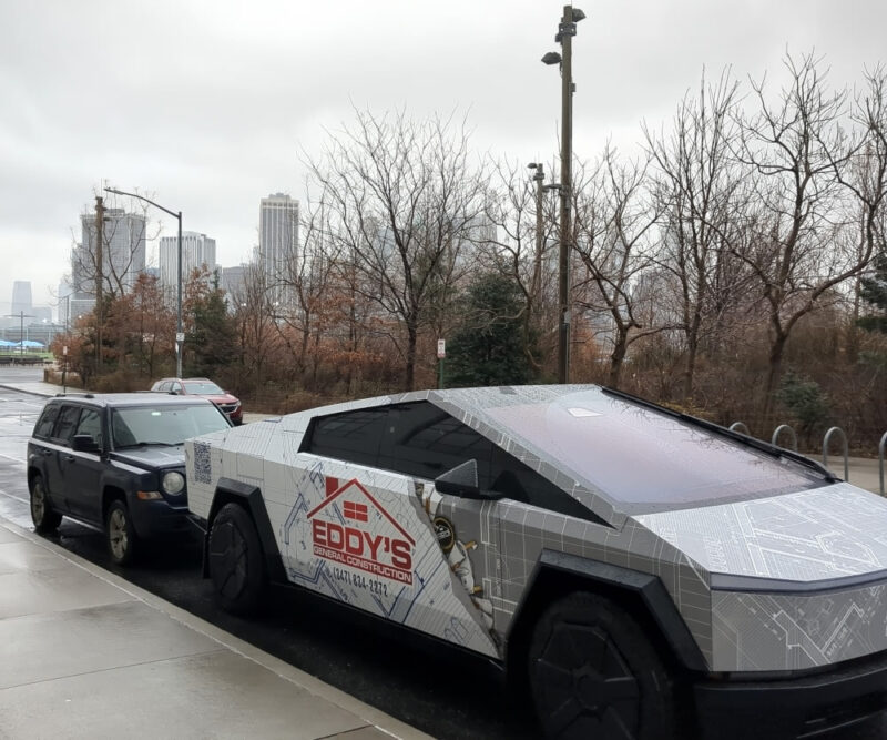 Photograph of a white and gray Cybertruck with a red Eddy's General Construction logo on its side parked at the Joralemon Street exit of Brooklyn Bridge Park. There is one car parked right next to the Cybertruck. In the background you can see baren trees and a gray, overcast sky, with the Manhattan Skyline across the East River in the distance.