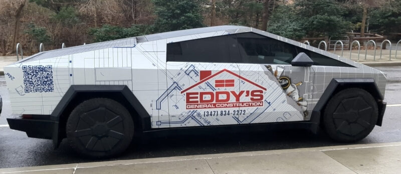 An Eddy's General Construction Cybertruck parked alongside the sidewalk at the Joralemon Street exit of Brooklyn Bridge Park. The car is mostly white with light grays and blues making up a blueprint pattern. The red Eddy's General Construction logo is on the side door.