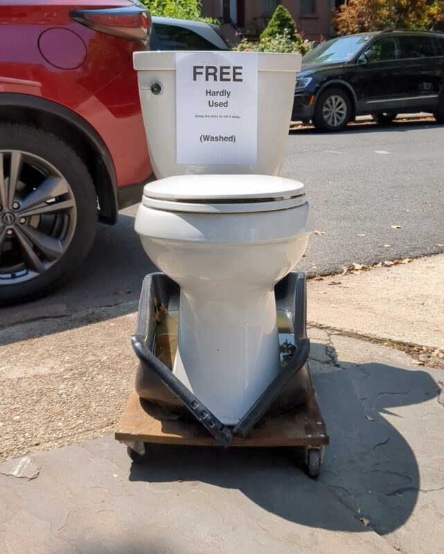 A toilet on a dolly outside on the curb in Carroll Gardens, Brooklyn. There is a piece of paper taped to the tank with typed sans serif font. It reads, from top to bottom: "FREE / Hardly Used / (Keep the dolly and roll it away) / (Washed)"