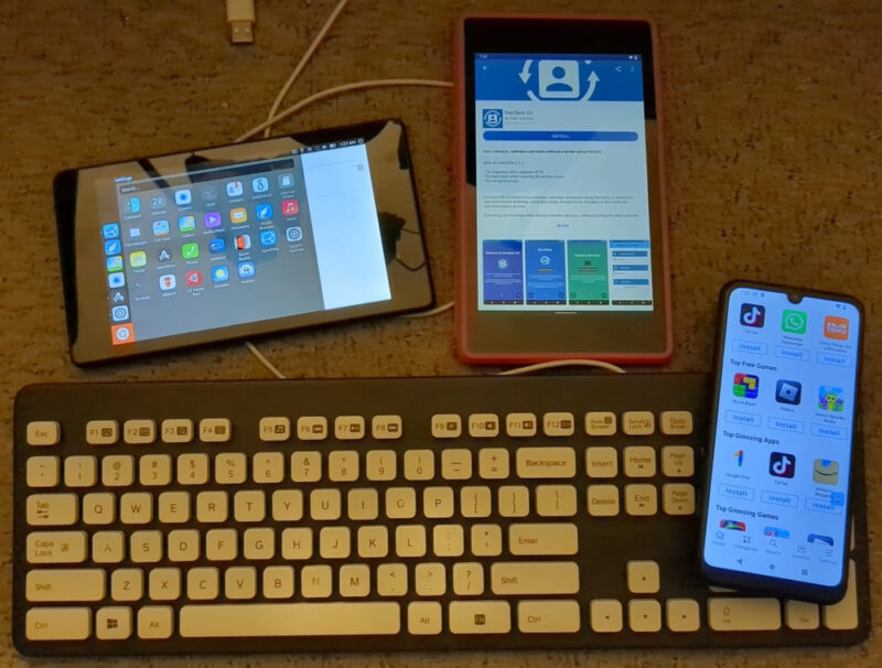 We see two Google Nexus 7 2013 tablets laid out next to each other above a keyboard and phone, all on a carpeted floor. The left tablet is open to the Ubuntu Touch launcher. The right tablet, running LineageOS, is open to F-Droid with the DecSync CC app in F-Droid. On the bottom, we have a gray and white Logitech K310 keyboard. Resting on the keyboard's numpad is a Teracube 2e phone open to the /e/ OS App Store.