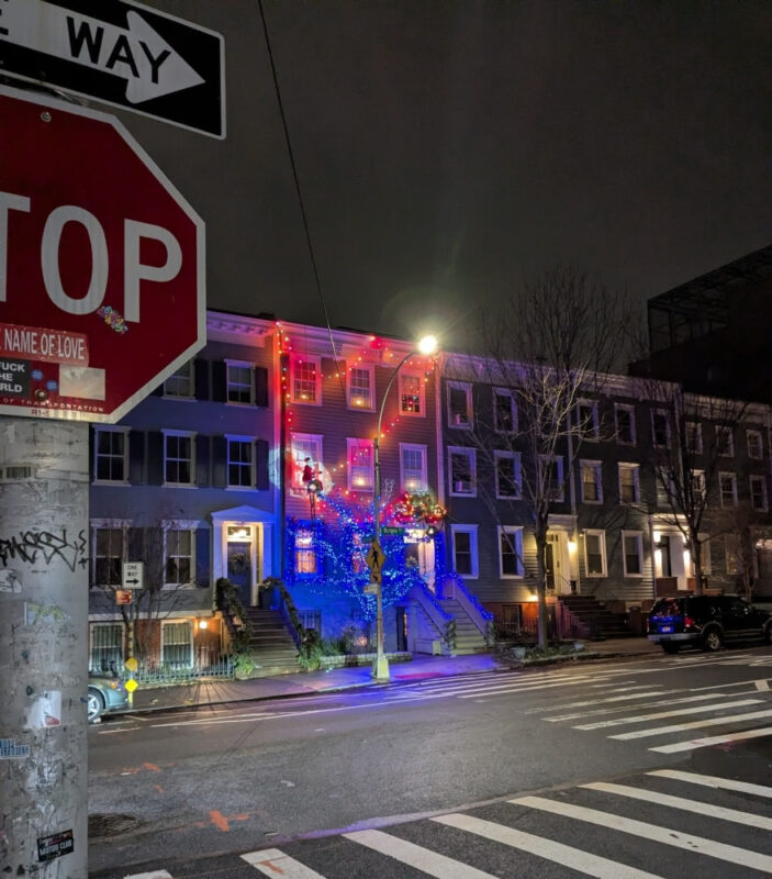 Photograph taken in the evening in Boerum Hill, Brooklyn, at the intersection of Bergen Street and Boerum Place. In the foreground there is a hybrid ONE WAY/STOP sign. Across the street we see red and blue Christmas lights on a building.