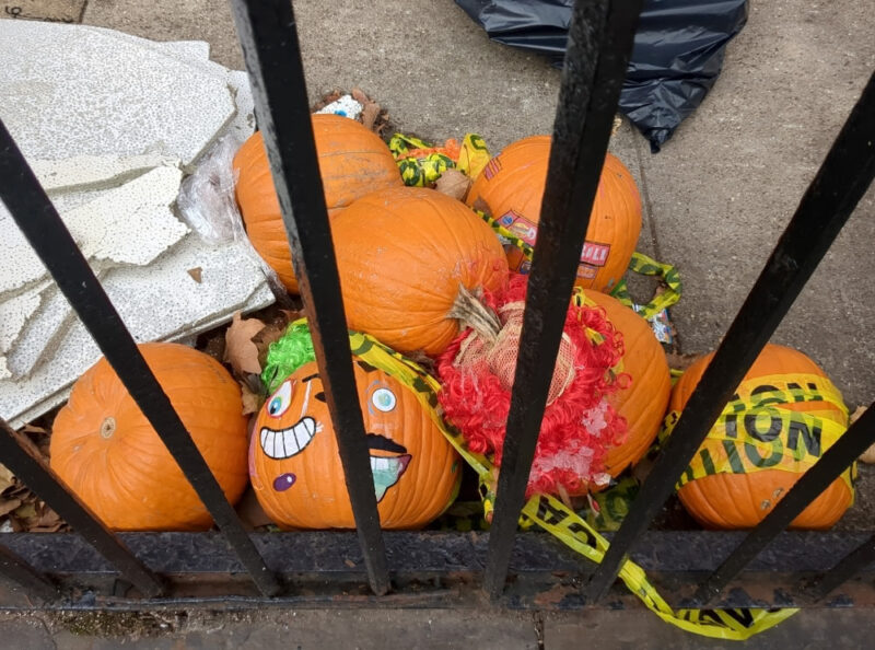 Seven decorative pumpkins piled on a sidewalk behind a cast iron fence. There is caution tape strewn about the pumpkins. One pumpkin has face stickers on it. Another had red tinsel on its crown. A third has stickers with the logo of a local deli.