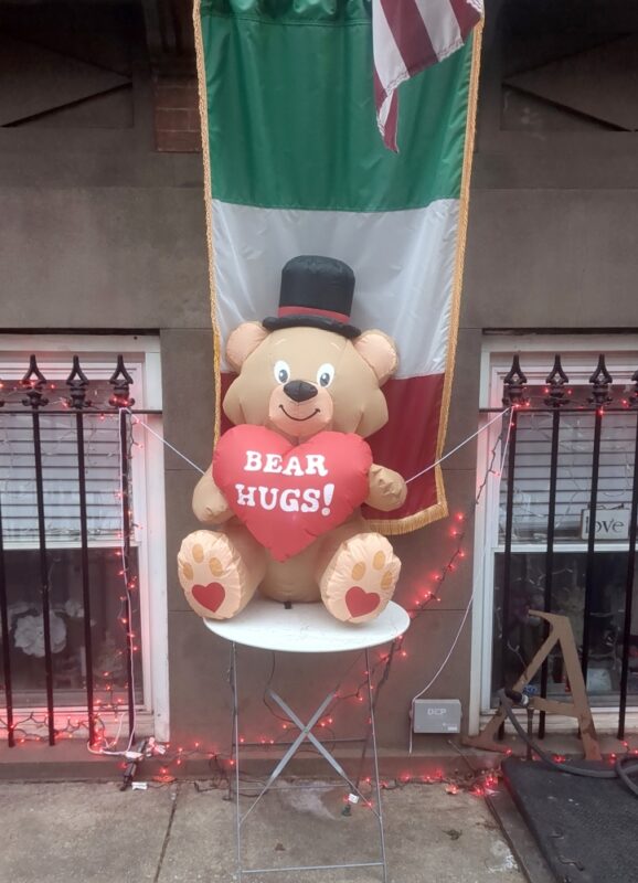 Inflatable brown bear in a tophat holding a red heart which says BEAR HUGS! and wearing an inflatable black tophat seen on a white folding table in front of a brownstone. There is an Italian flag behind the bear and a U.S. flag right above.