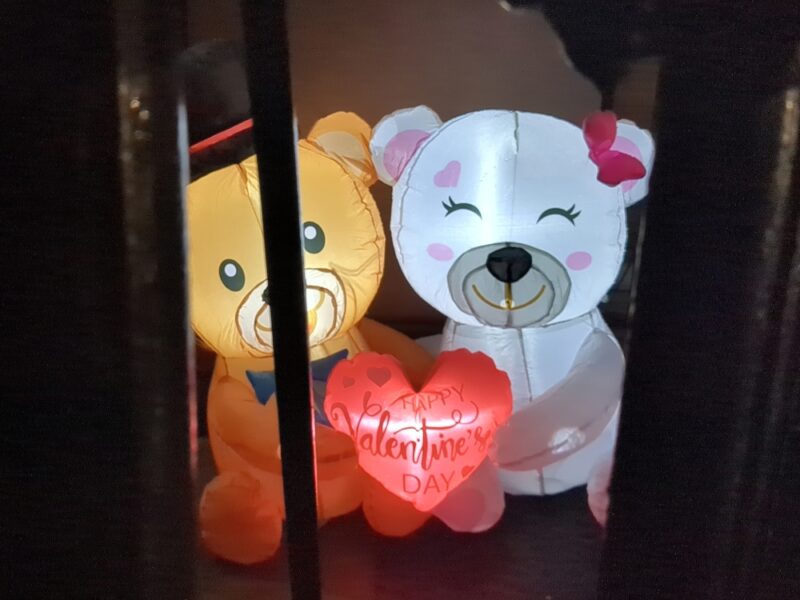 Inflatable Valentine's Day decoration featuring a small brown bear with a black tophat on the left and a white bear with a pink bow on the right. The brown bear is somewhat obscured by a black cast iron fence. They are smiling while holding a heart which says Happy Valentine's Day.