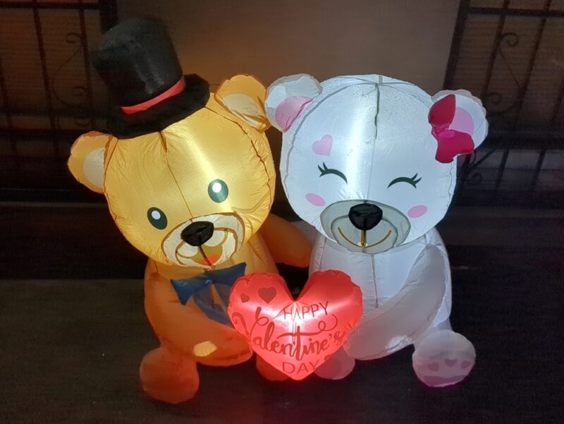 Inflatable Valentine's Day decoration featuring a small brown bear with a black tophat on the left and a white bear with a pink bow on the right. They are smiling while holding a heart which says Happy Valentine's Day.