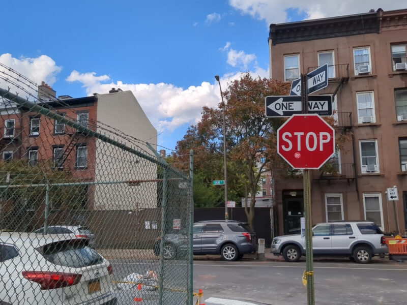 Photograph of a hybrid Stop-One Way sign on Kane Street in Brooklyn with a fenced-off lot to the left and small apartment buildings ahead.